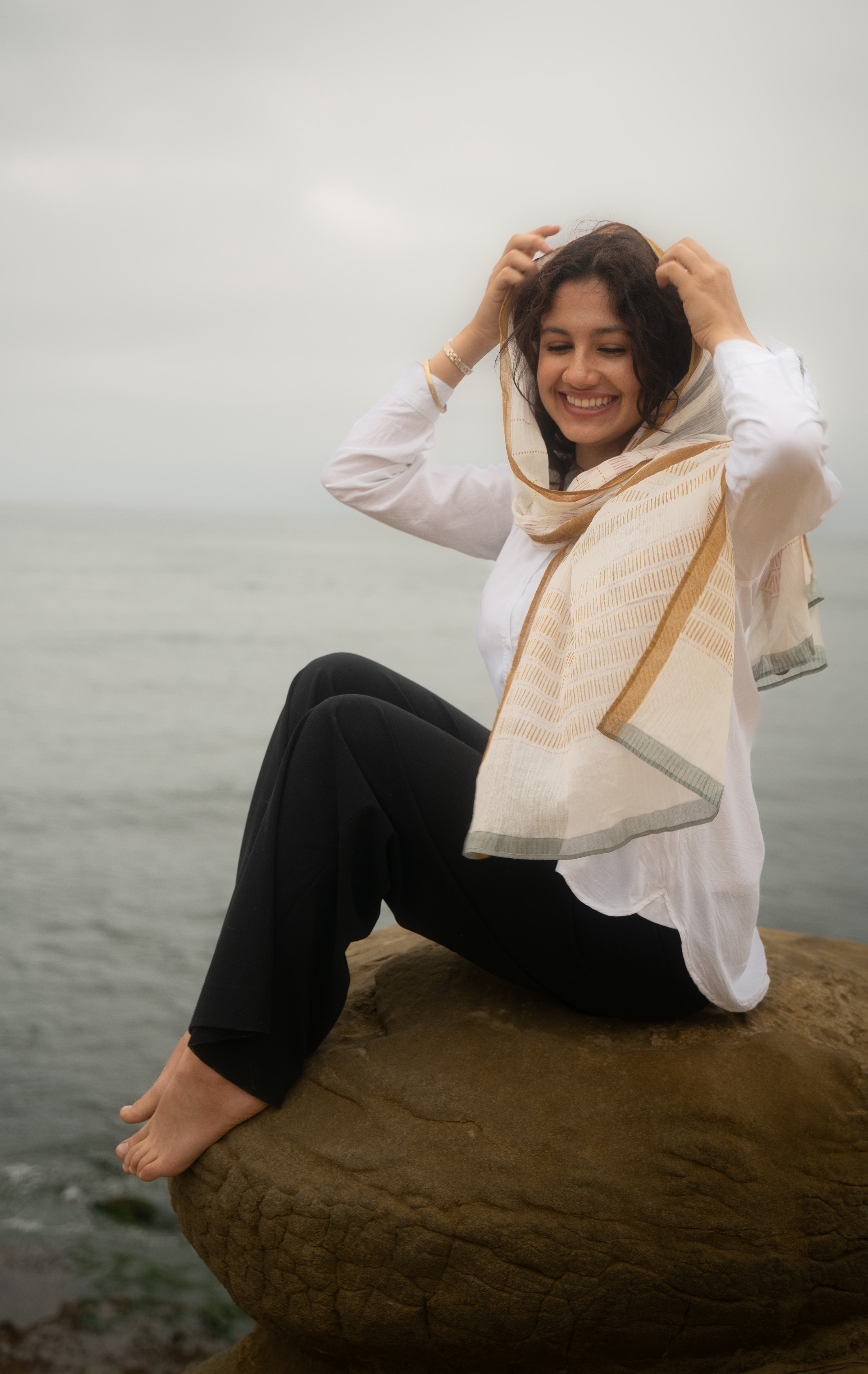 Woman sitting on a rock by a body of water wearing a white shirt and black pants with a beige scarf.